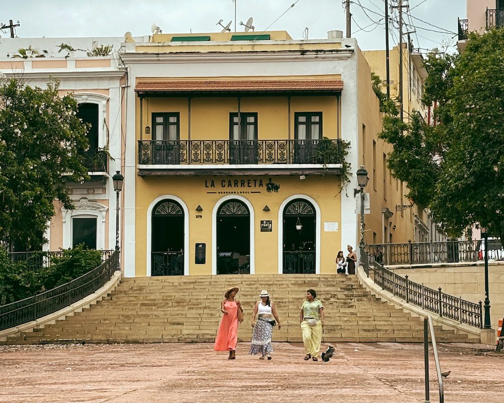 A yellow building by the Process Promenade in Old San Juan with three women in colorful clothing in front.